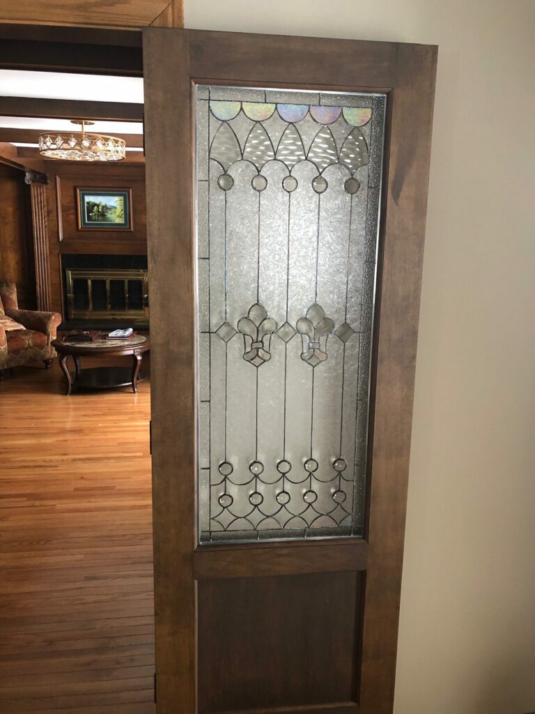 A rectangular leaded stained glass door panel with clear textured and beveled glass, featuring three prominent fleur-de-lis motifs, iridescent scalloped glass at the top, and clear circular accents, set within a dark wooden interior door.