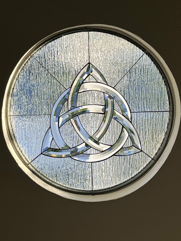 A circular stained glass window featuring a prominent central clear beveled Triquetra Celtic knot design. The knot is surrounded by clear, heavily textured (likely glue chip) glass, all held together with black leading lines and set within a white frame.