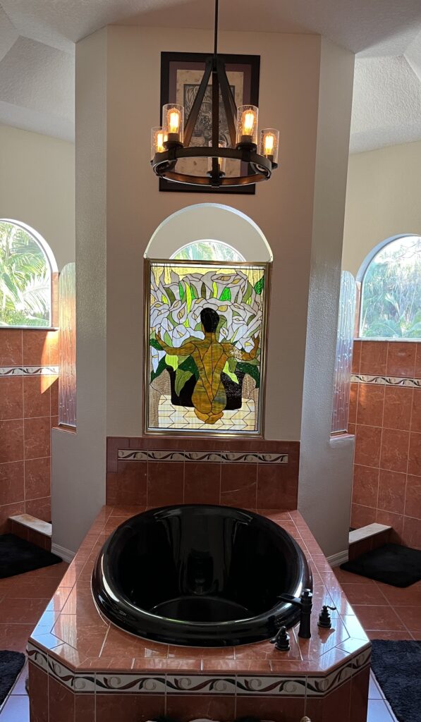 A rectangular leaded stained glass panel with an arched top and a golden frame, depicting a stylized woman from the back surrounded by large white calla lilies and green foliage. The panel is installed in a bathroom wall above a black oval soaking tub, with terracotta-colored tiled walls and arched windows visible on either side.