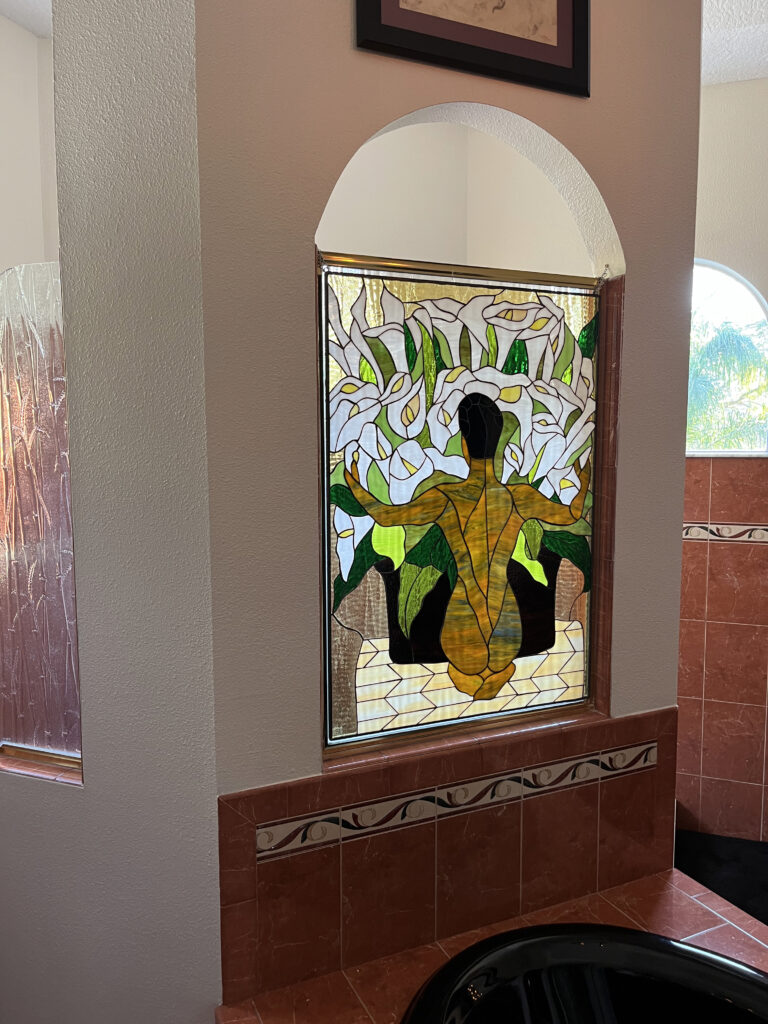 A large rectangular stained glass panel installed in a bathroom's arched opening, depicting the bare back of a person (golden and brown glass) surrounded by numerous white calla lilies, green leaves, and textured golden background glass. Reddish-brown tiled walls and a black bathtub are visible below and to the side.