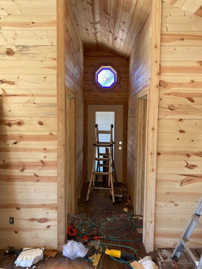 An octagonal stained glass window features a vibrant blue glass border framing an intricate clear, textured, and subtly colored geometric medallion in its center. The window is installed high in a narrow hallway with light pine wood paneling on the walls and angled ceiling. Construction tools and a wooden ladder are visible on the floor below, indicating the space is under renovation.