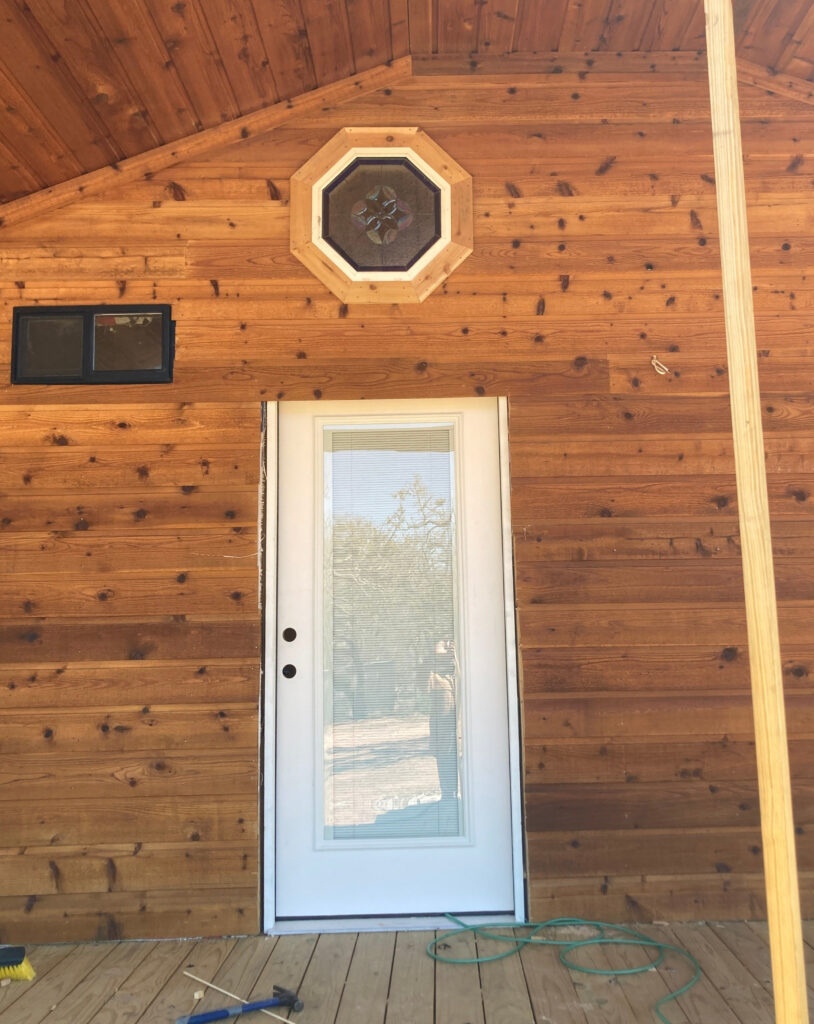 An octagonal insulated stained glass window, set within a light wood trim, featuring a vibrant leaded geometric floral medallion in shades of blue, purple, and amber. It is installed high on a rustic knotty pine exterior wall above a white full-lite entry door, depicting a building under construction.