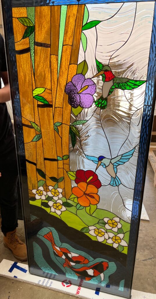 Rectangular stained glass panel depicting a vibrant tropical scene with golden bamboo stalks, two colorful hummingbirds (red and blue), various flowers, and two orange and black koi fish swimming in a pond.