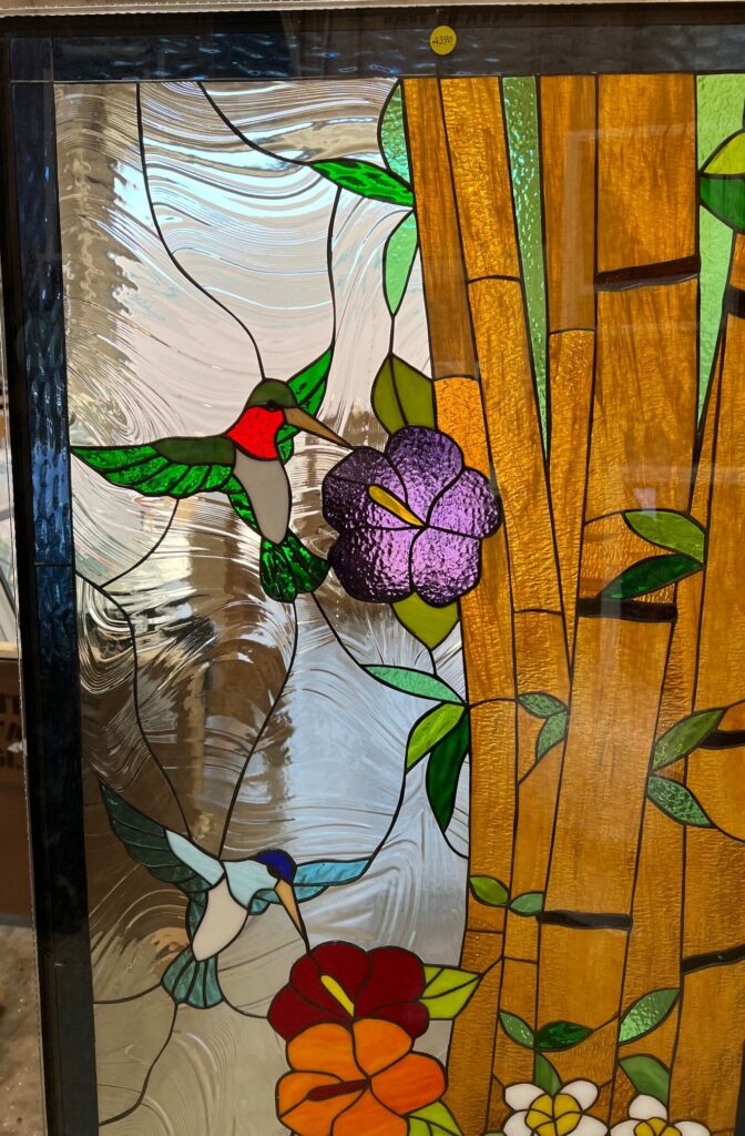 Rectangular stained glass panel depicting two colorful hummingbirds, one red-throated and one blue, feeding from vibrant purple, red, orange, and white flowers. Golden bamboo stalks and green leaves frame the scene against a clear, swirling textured glass background.