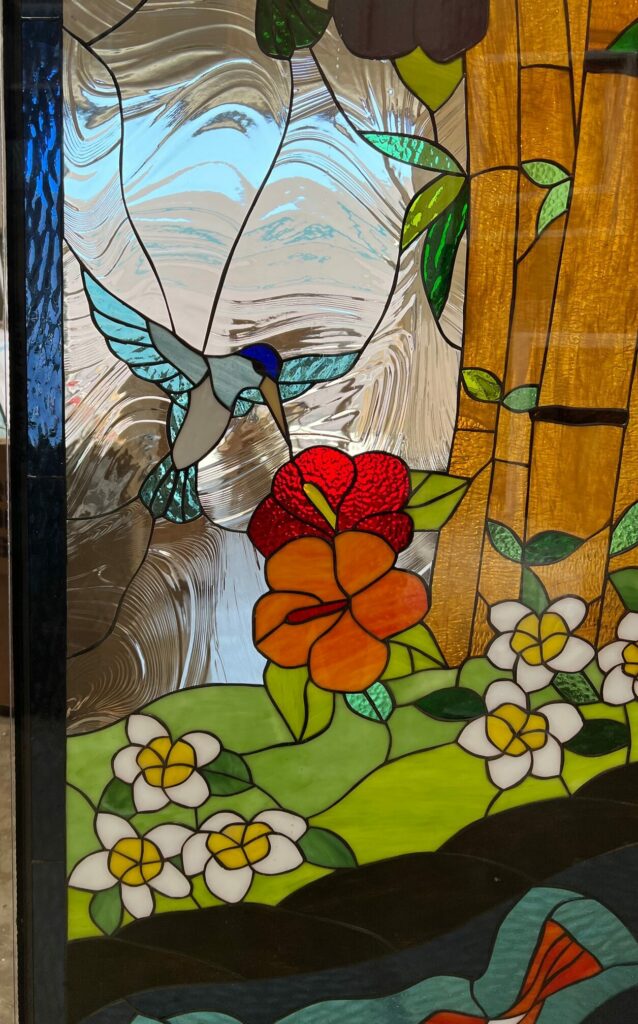 A vibrant leaded stained glass panel depicting a blue hummingbird hovering near bright red and orange hibiscus flowers, surrounded by golden bamboo stalks, green foliage, and white flowers, all against a clear, heavily textured wavy glass background.