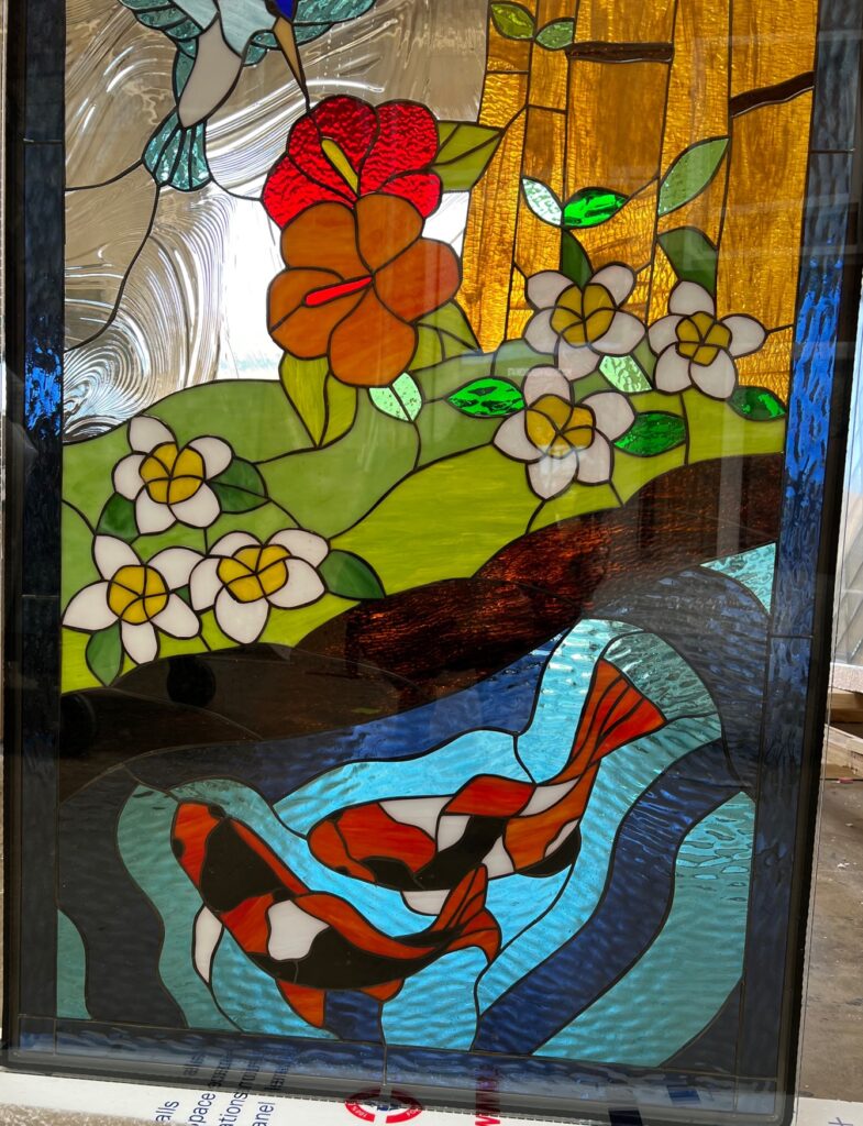 A rectangular leaded stained glass panel depicting a vibrant nature scene. A blue and white hummingbird hovers over red and orange hibiscus flowers and white flowers with yellow centers. Two orange, black, and white koi fish swim in a blue and turquoise pond below, with golden glass representing foliage in the background.