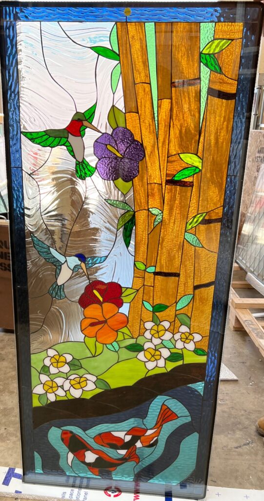 A rectangular stained glass panel depicting a vibrant tropical scene. Golden bamboo stalks rise on the right, while two colorful hummingbirds, one red-headed and one blue-headed, fly near purple, red, and white hibiscus-like flowers. Below, a pond features two orange, black, and white koi fish swimming in blue and green water. The background is clear, swirling textured glass, and the panel has a dark frame.