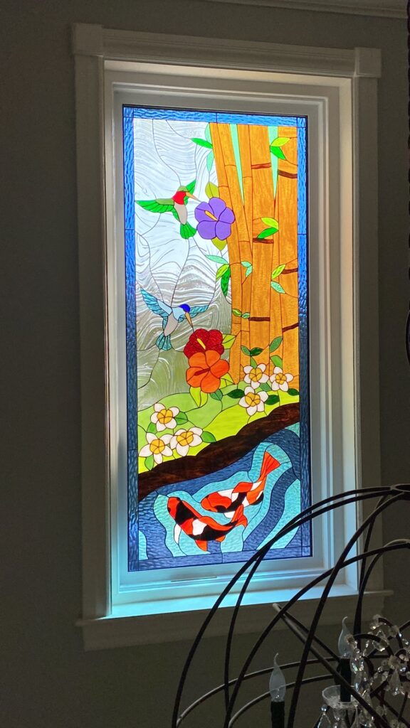 A rectangular stained glass window panel depicting a vibrant tropical scene with two hummingbirds, colorful flowers, golden bamboo stalks, and two koi fish swimming in blue water, all set against a textured clear glass sky.