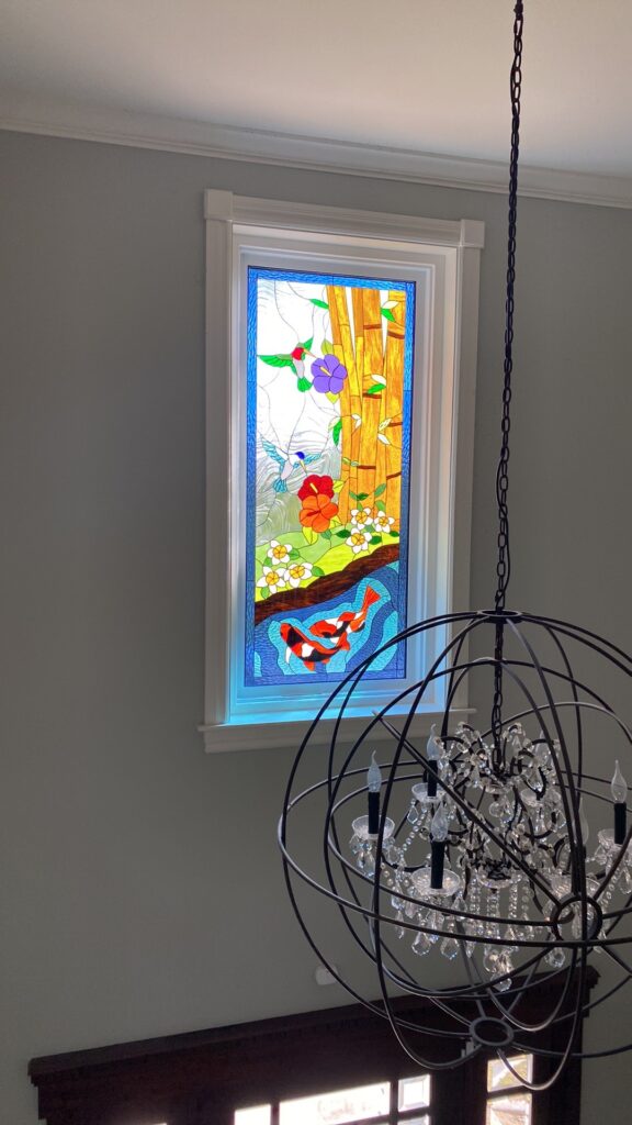 A rectangular stained glass window panel depicting a vibrant natural scene with golden bamboo, a green hummingbird near a purple flower, a blue hummingbird, red and white blossoms, and two orange and black koi fish in blue water.