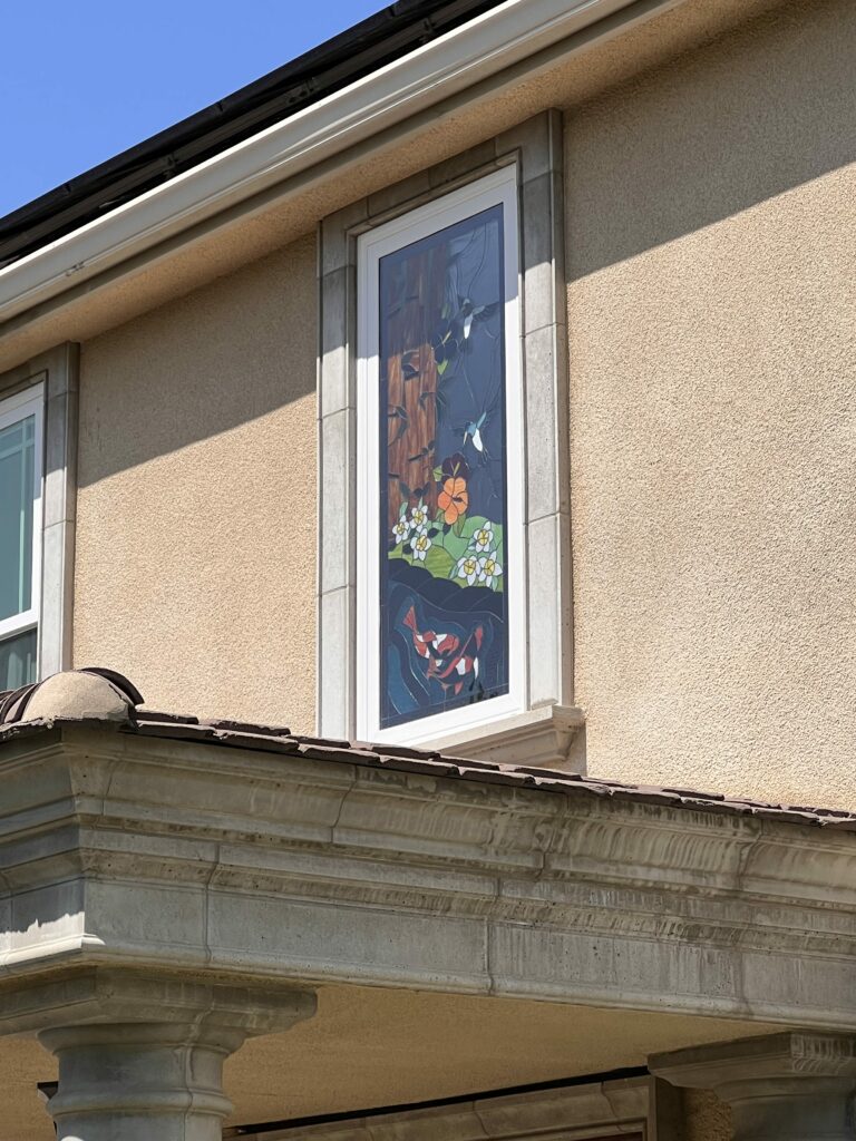 A tall, rectangular stained glass window panel installed in a white-framed window on a beige stucco house. The artwork depicts a vibrant nature scene with two white hummingbirds in flight, colorful white and orange flowers with green leaves, and stylized red and white koi fish swimming in dark blue water.