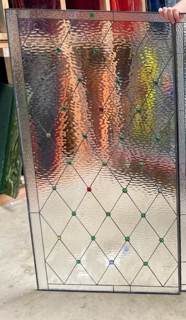 A rectangular leaded stained glass panel featuring a clear diamond lattice pattern. The design incorporates both rippled (water glass) and smooth clear glass, with small, round, multi-colored glass jewels in green, red, yellow, and blue embedded at many of the diamond intersections. The lead lines are dark.
