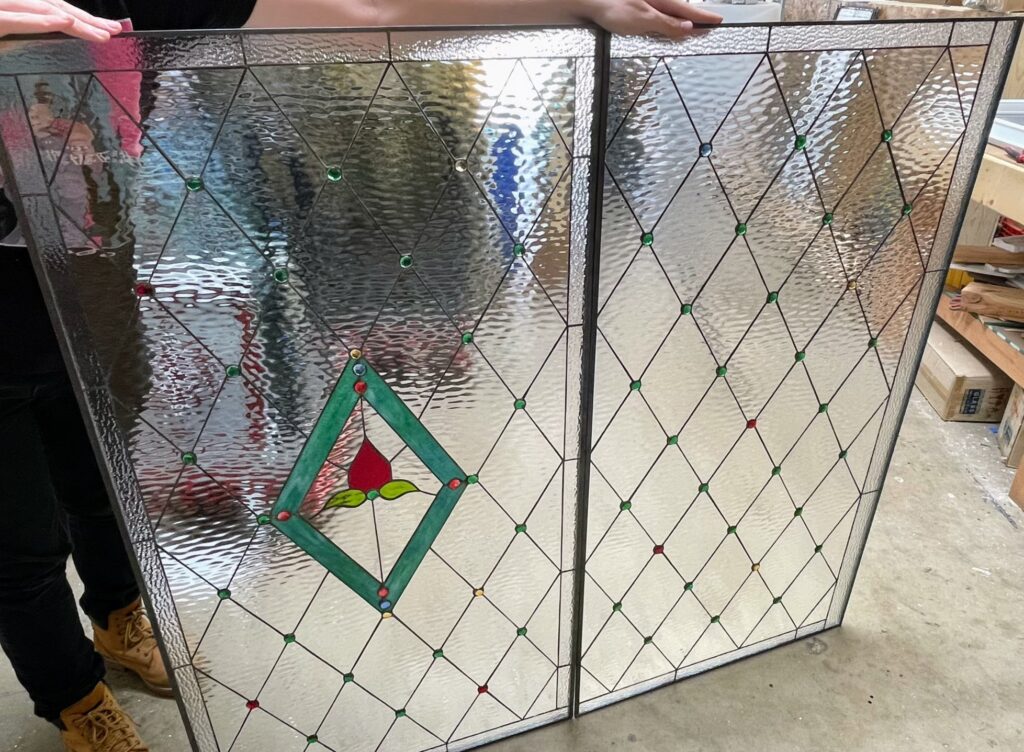 Two rectangular stained glass panels held by a person. Each panel displays a diamond grid pattern created with clear, ripple-textured glass and dark lead lines. Small, round glass jewels in green, red, blue, and yellow are interspersed at the diamond intersections. The left panel features a central emerald green diamond frame encompassing a red flower bud with green leaves.