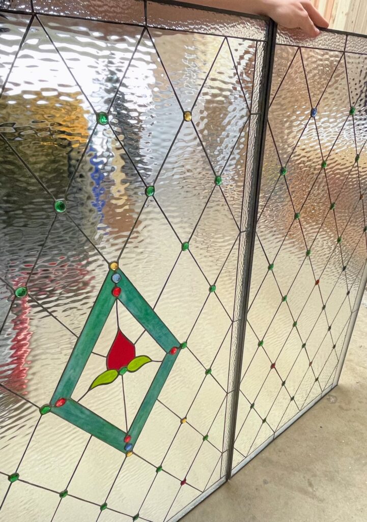 Two rectangular leaded stained glass panels, featuring clear heavily textured glass in a diamond grid pattern. Small, round green, red, yellow, and blue glass jewels are embedded at many lead intersections. The left panel includes a central green diamond frame enclosing a stylized red rosebud with green leaves.