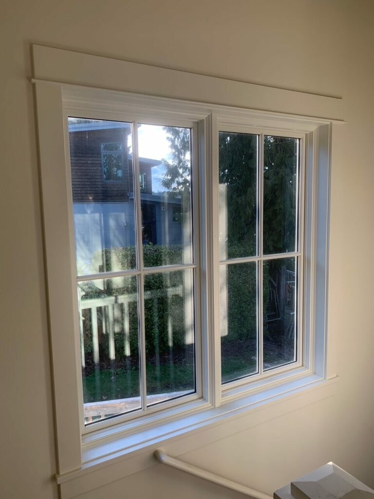 A well-lit interior shot of a white-framed double-hung window with clear glass panes and muntins, looking out to a suburban backyard with a house and green trees.