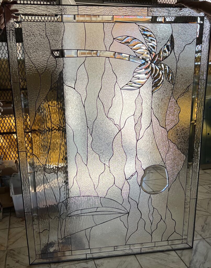 A rectangular leaded stained glass panel featuring a stylized horizontal palm tree with clear beveled fronds and trunk in the upper right. The background consists of various clear, textured glass pieces in flowing, organic patterns, evoking water or sky. In the lower left, a stylized fish or marine life figure is discernible, crafted with clear beveled details, alongside a prominent circular beveled glass piece in the lower right. The entire panel is crafted from clear, textured, and beveled glass, providing privacy and exquisite light refraction.