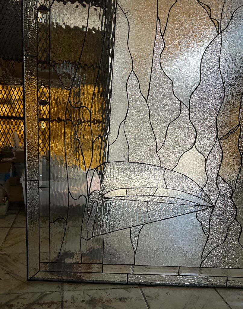 A rectangular leaded stained glass panel with an abstract, organic design. It features various clear textured glass types, including a large, prominent, curving leaf or wing-like form, all connected by dark lead lines. The panel reflects warm, blurred light from the background, hinting at a workshop setting.