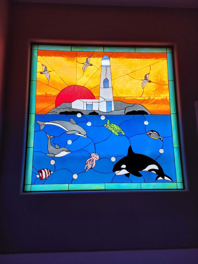 A rectangular stained glass panel depicting a vibrant marine and coastal scene. The upper half features a bright orange and yellow sky with a textured red sun, a white lighthouse on a rocky island, and three grey seagulls flying. The lower half is a deep blue ocean populated by two grey and white dolphins, a green sea turtle, a red and white striped fish, a smaller dark fish, a pink jellyfish, and a prominent black and white orca, with white bubbles scattered throughout.