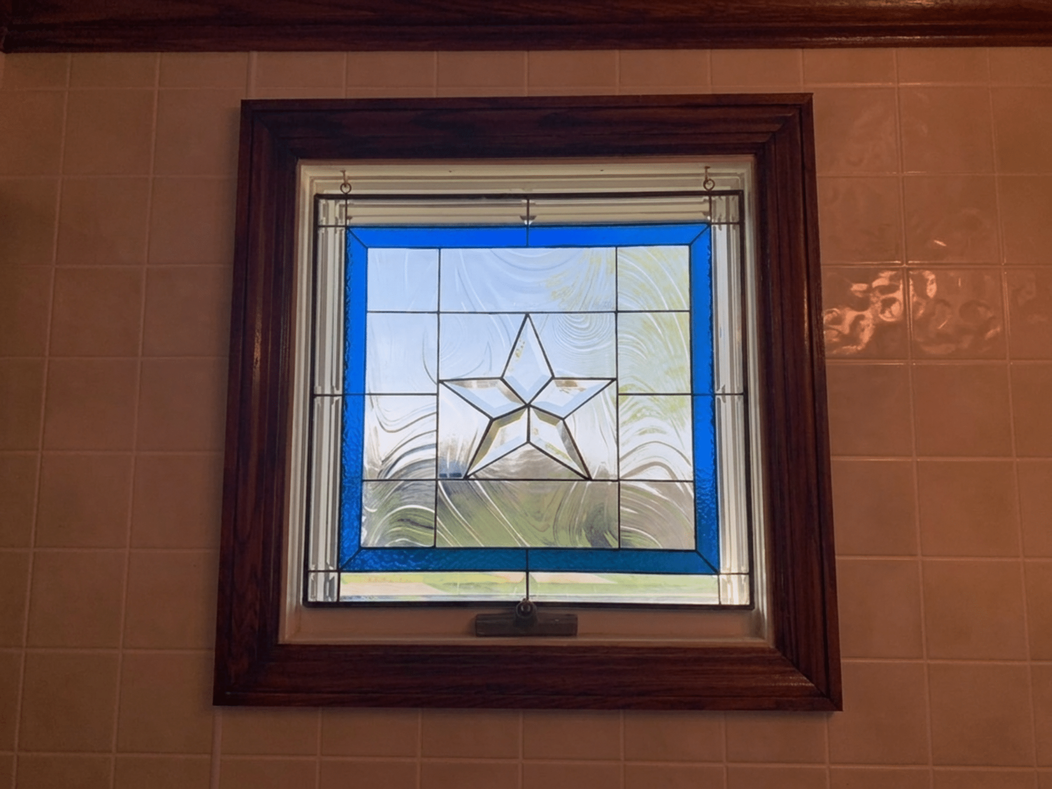 A square stained glass panel featuring a central beveled star, framed by a vibrant blue border, set within a dark wooden window frame against a light-tiled wall.
