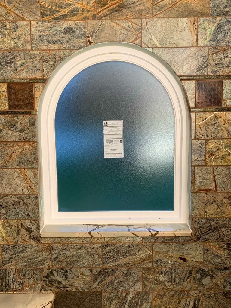 A white-framed arched window with textured, obscure privacy glass, set within a wall of rectangular dark green and brown marble-like tiles with golden veins. Two square hammered copper accent tiles are visible to the left and right sides of the arch. A white NFRC energy performance label is affixed to the lower center of the glass.