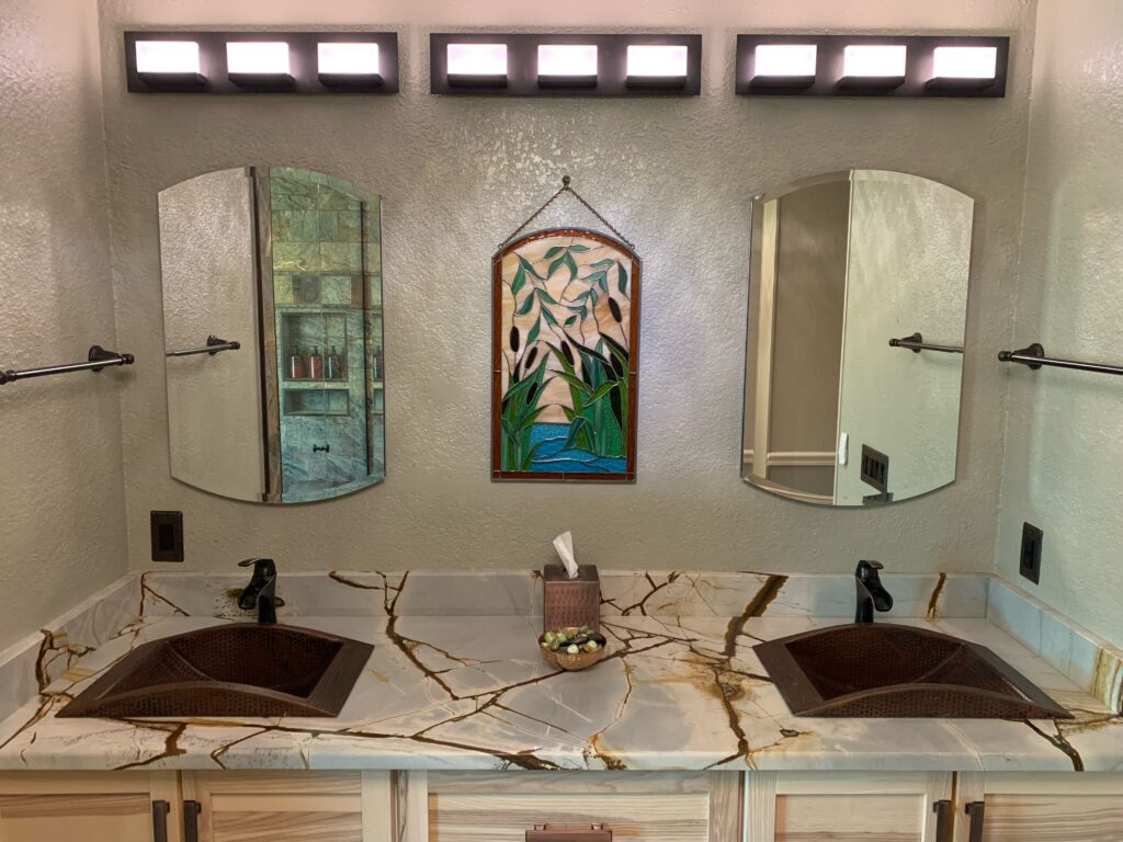 A rectangular stained glass panel with an arched top, featuring a wetland scene of tall green cattails, marsh foliage, and blue water, hanging by a chain between two mirrors in a bathroom.