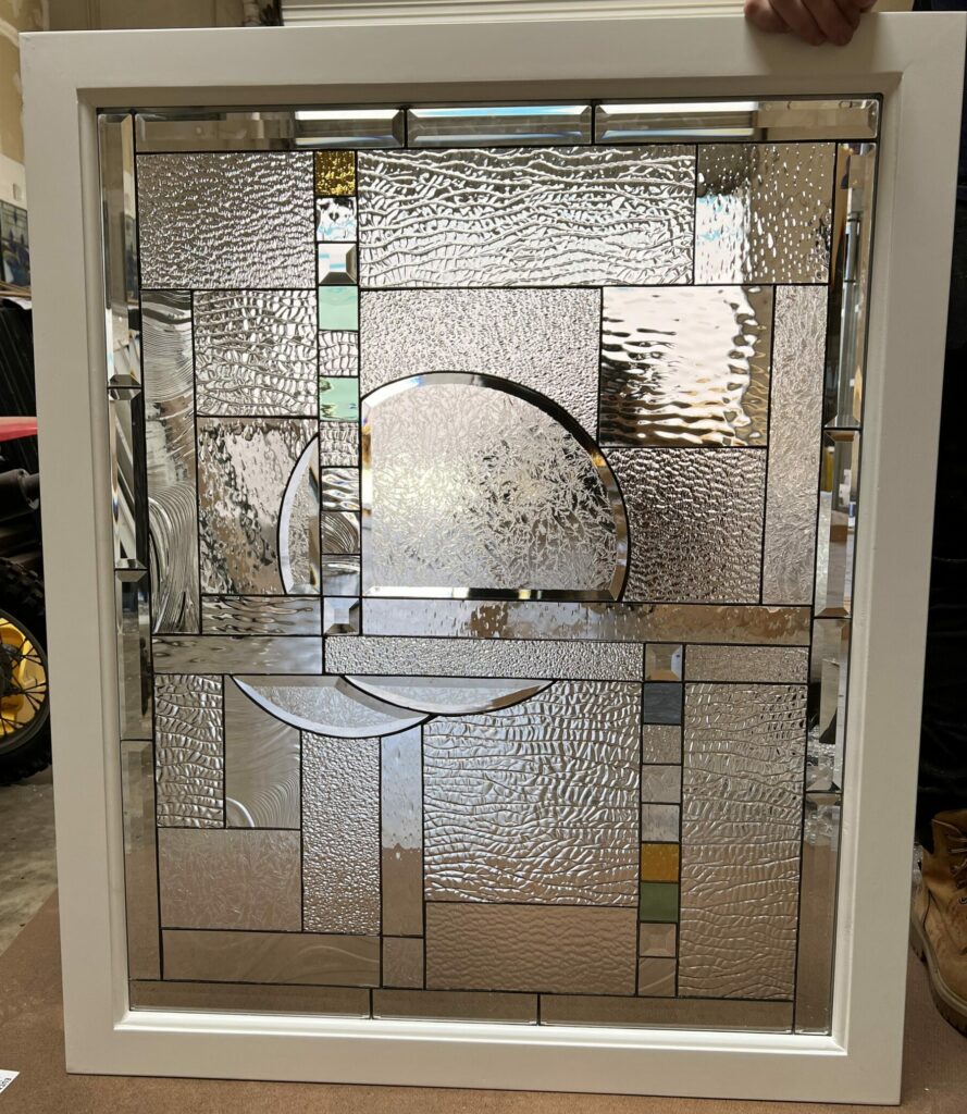 A rectangular leaded glass window panel, set in a white frame, showcasing a modern abstract geometric design. It features diverse clear textured glass, including large central circular and semi-circular patterns, various patterned clear panels, and small accent squares of amber, teal, and light green. Beveled glass pieces are incorporated along the edges.