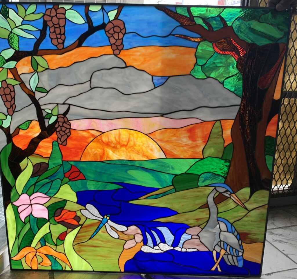 A rectangular stained glass panel depicting a vibrant nature scene. A sunset with orange and yellow hues illuminates a blue river, where a gray heron stands and a blue dragonfly hovers. Lush green trees with brown pinecones and colorful flowers frame the river.