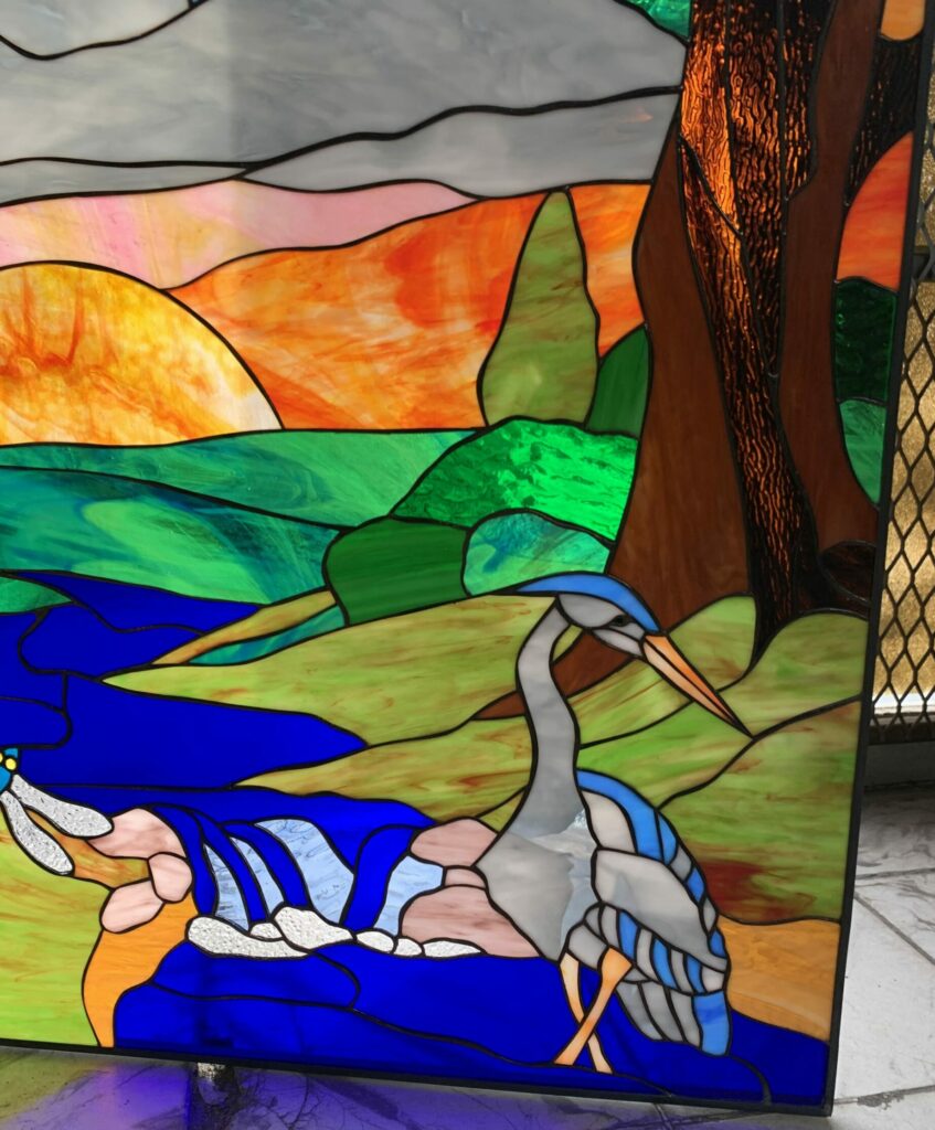 A rectangular leaded stained glass panel depicting a majestic blue and grey heron wading in deep blue water. The background features a vivid sunset landscape with glowing orange and green rolling hills, a tall brown tree trunk, and a light grey sky.