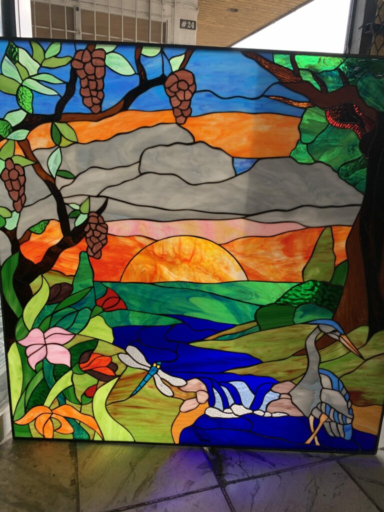 A rectangular stained glass panel depicting a vibrant natural landscape at sunset. In the foreground, lush green foliage and colorful flowers frame a deep blue river with a small waterfall. A blue dragonfly hovers near the water, and a grey and blue heron stands on the right bank. Tree branches with brown pine cone-like clusters extend from the top left and right. The background features an orange and red sunset over distant hills and a blue sky above, with grey clouds.