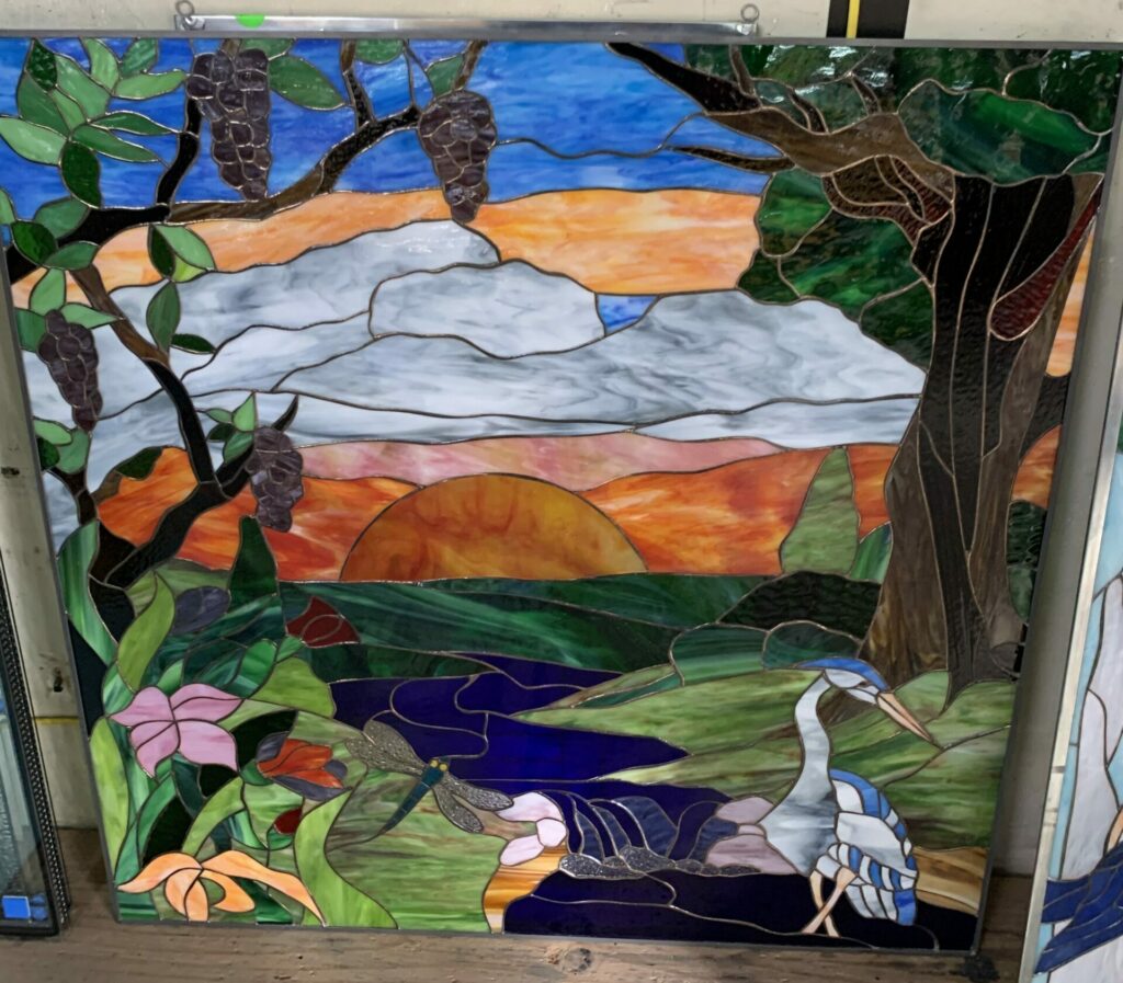 A rectangular stained glass panel depicting a vibrant nature scene at sunset. A bright orange and red sun sets over a distant horizon, beneath a blue sky with white clouds. In the midground, rolling green hills lead to a deep blue river. On the left, lush green branches with clusters of dark purple grapes frame the view. On the right, a large dark tree trunk with green foliage creates a natural border. In the lower left, delicate pink and orange wildflowers bloom, alongside a detailed dragonfly with metallic wings. In the lower right, a graceful blue and white heron stands in the water. The panel uses varied textured and colored art glass pieces joined by copper foil, set within a metal frame.