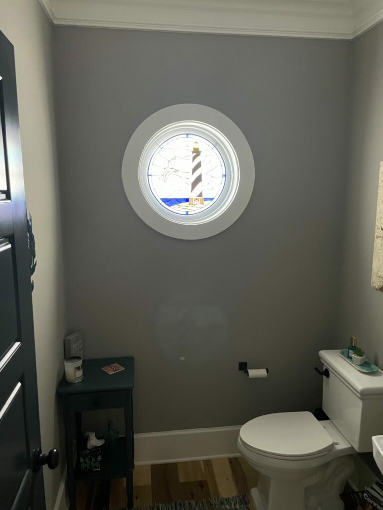 A round stained glass window featuring a black and white striped lighthouse with a yellow light, a white seagull flying, a sky with clear textured glass, and blue water with a tan shoreline. The window is set within a white circular frame in a gray wall.