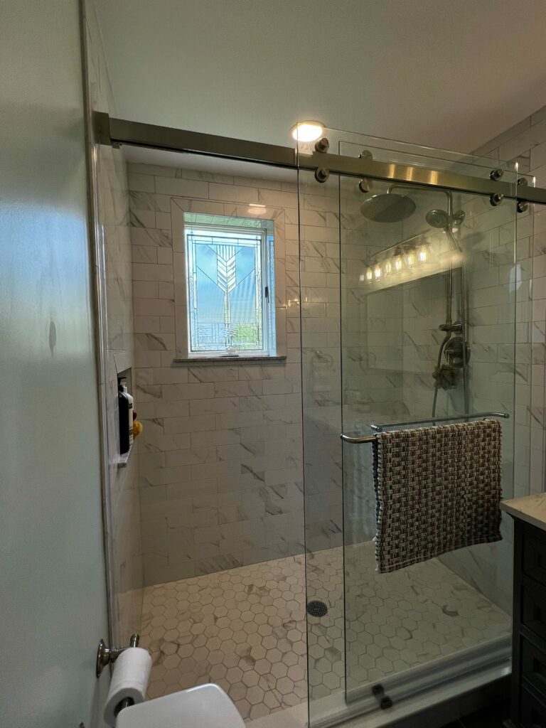 A rectangular stained glass window with a clear, geometric Art Deco-inspired design, featuring textured and possibly beveled glass segments, installed in a modern tiled shower stall with white subway and hexagonal floor tiles. The window brings natural light into the shower while providing privacy.