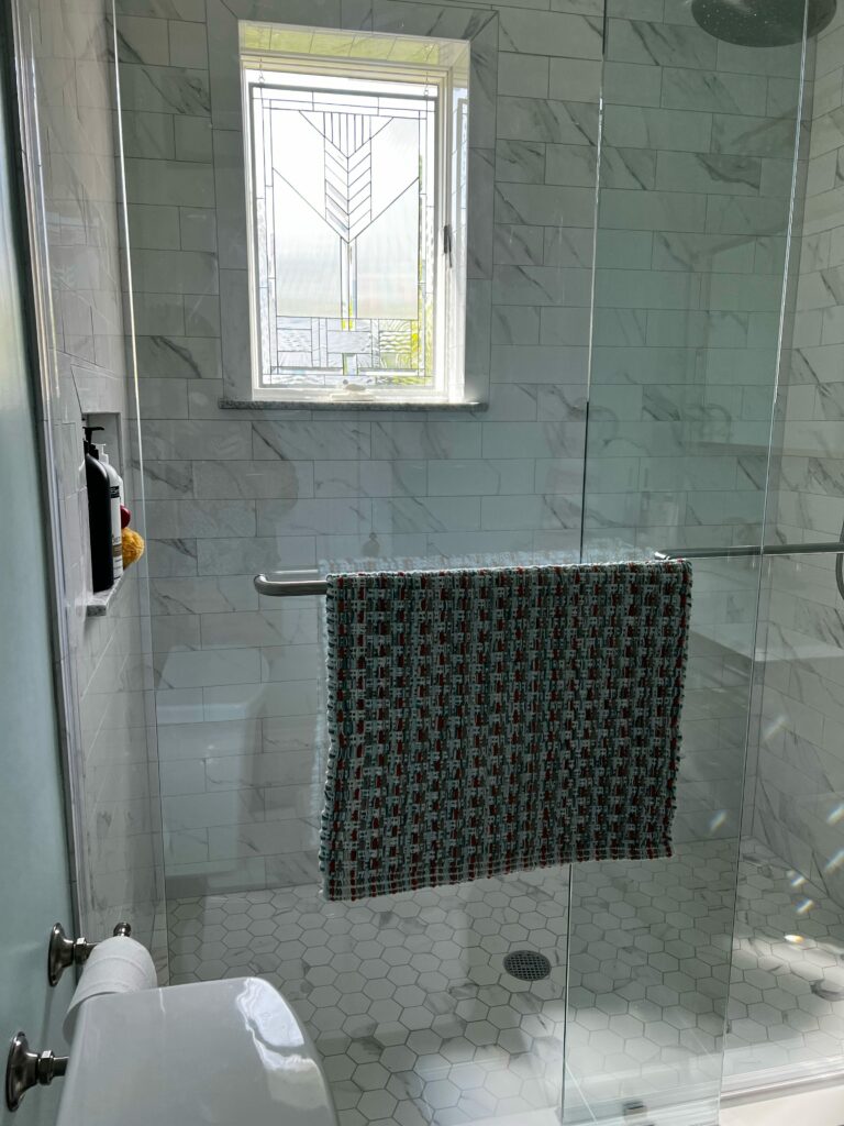 A rectangular leaded stained glass transom window featuring an Art Deco-inspired geometric pattern with various clear textured and subtly beveled glass panels, illuminating a modern bathroom shower with white subway tiles and hexagonal floor tiles.
