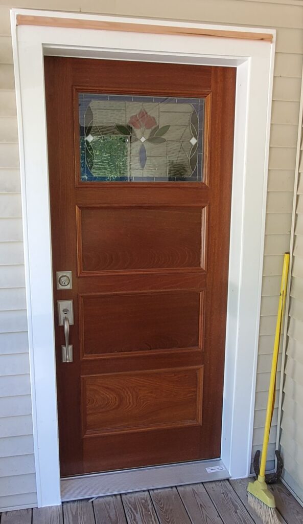 A rectangular leaded stained glass panel in a dark brown wooden entry door. The glass features a central stylized pink and red flower with green leaves and blue accents, surrounded by clear textured and beveled glass pieces. The door is set within a white frame on a porch.