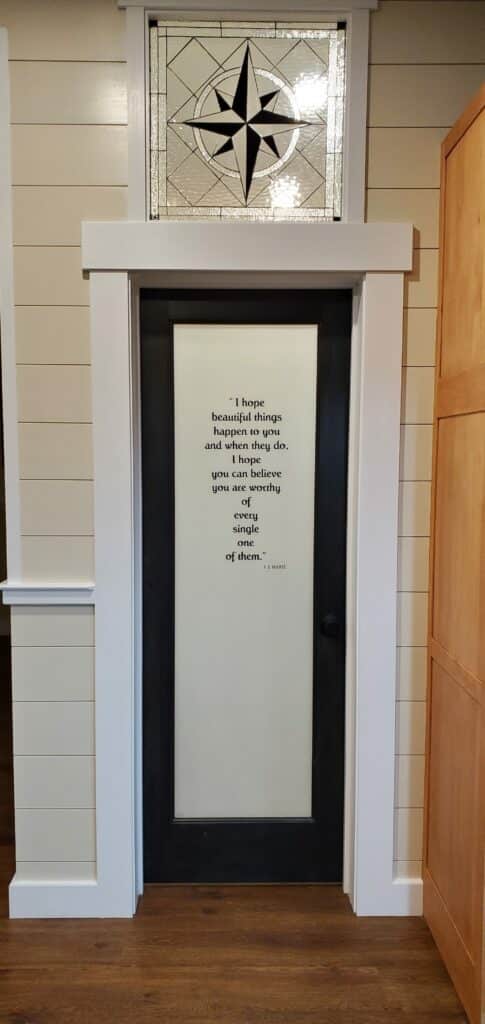 A rectangular leaded stained glass transom panel featuring a central black and clear compass rose design, surrounded by clear textured geometric glasswork, installed above a black interior door with a frosted glass panel.
