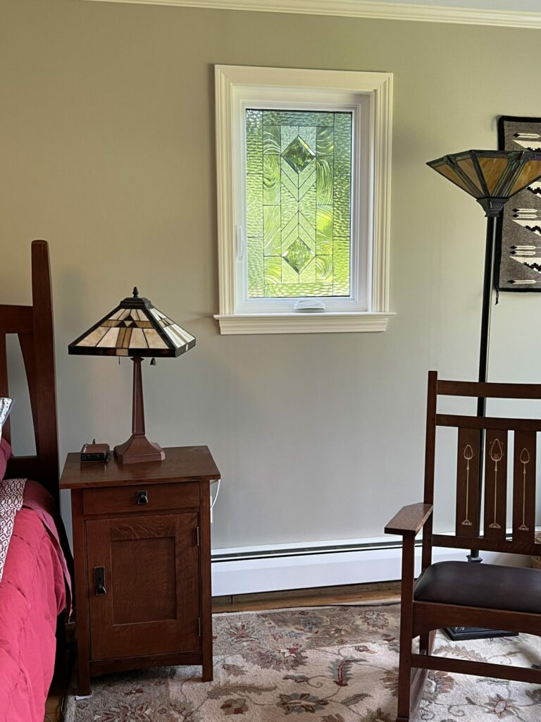 A rectangular, white-framed insulated window featuring a clear, textured, and beveled geometric stained glass panel, providing privacy while allowing natural light. The window is positioned above a Mission-style nightstand with a Tiffany-style lamp, in a room with light gray-green walls.