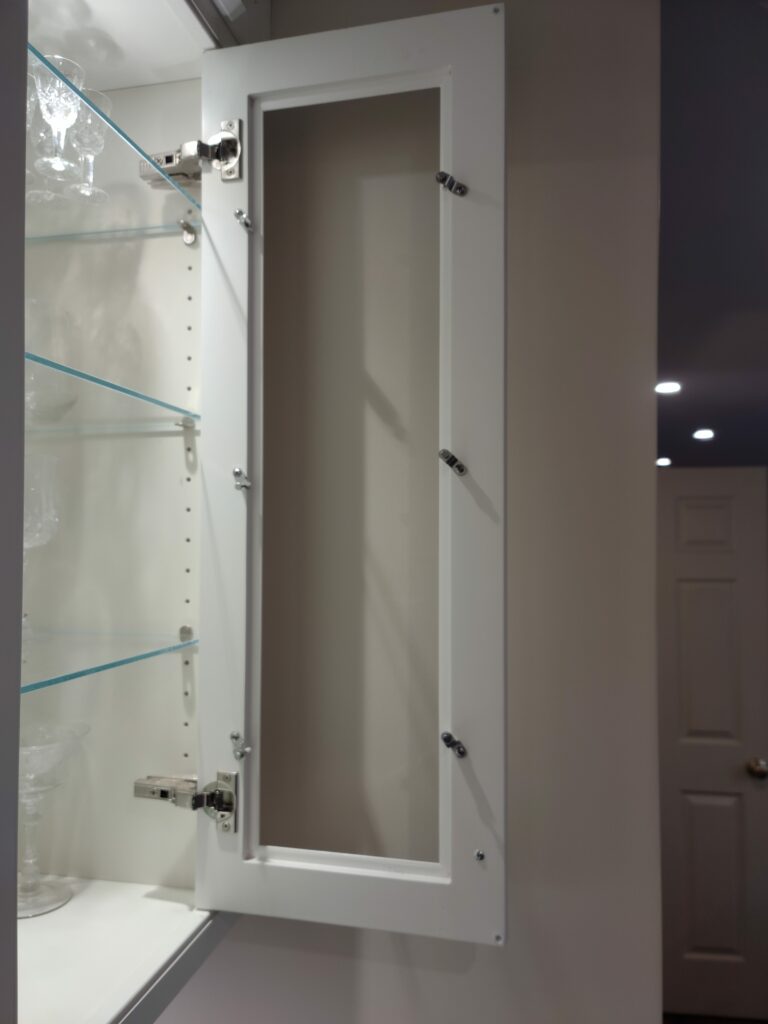 An open, white shaker-style cabinet door frame, empty and ready for a glass insert, revealing clear glass shelves and crystal stemware inside a cabinet.