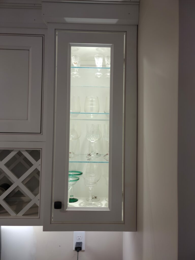 A tall, narrow white shaker-style kitchen cabinet with a clear glass panel door and a black handle, illuminated from within to display various stemware and drinking glasses on clear glass shelves. A white wine rack and another cabinet door are partially visible to the left, all against a light wall.