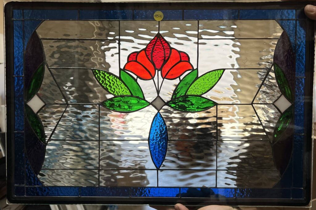 A rectangular leaded stained glass panel held by hands, featuring a central symmetrical Art Nouveau floral design with vibrant red petals, green textured leaves, and a blue stem-like element. The background consists of clear ripple-textured glass, framed by a dark blue glass border.