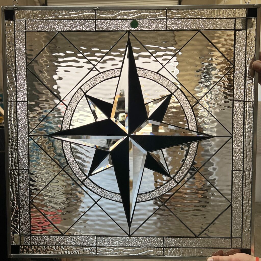 A square leaded stained glass panel featuring a prominent central compass rose. The compass points are crafted from clear beveled glass with dark opaque sections, encircled by a textured clear glass band. The surrounding field features a mosaic of clear textured glass, including wavy and stippled patterns, with a small green glass accent at the top.