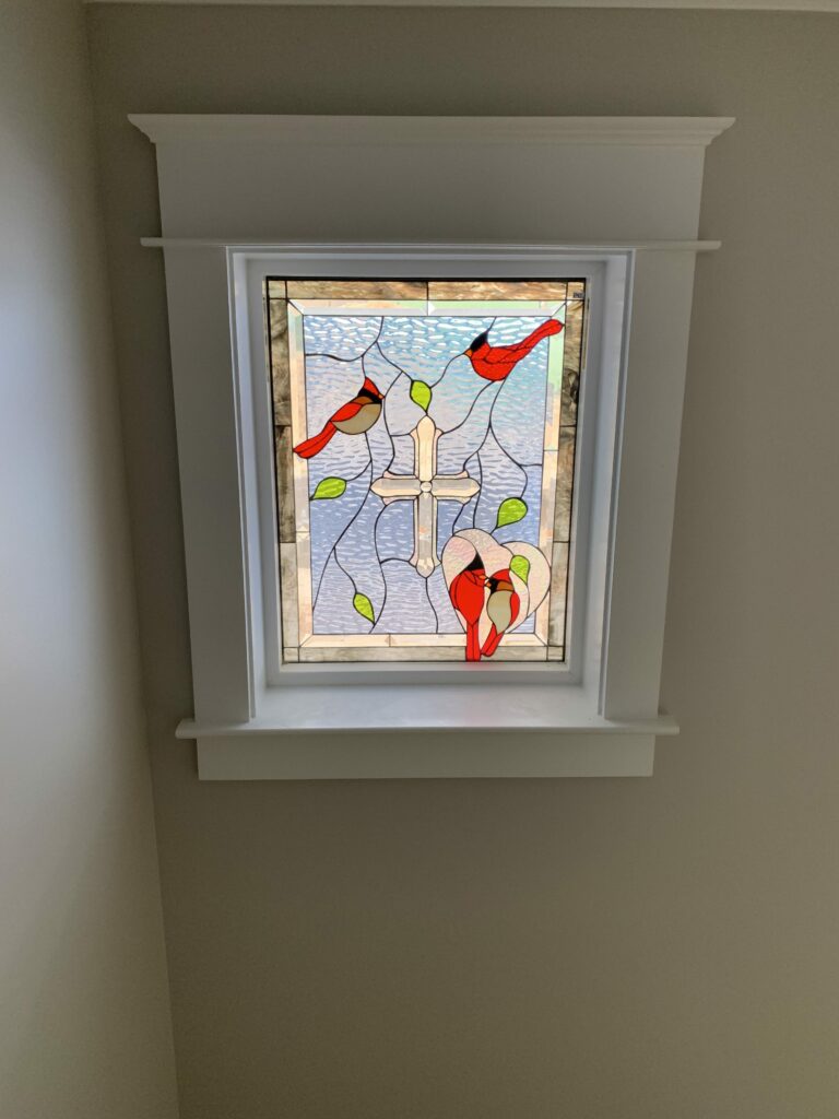 A rectangular leaded stained glass window panel featuring three vibrant red cardinal birds, green leaves, and a central white cross, set against a light blue rippled background and framed by clear textured and subtly beveled glass. The panel is installed in a white window frame.