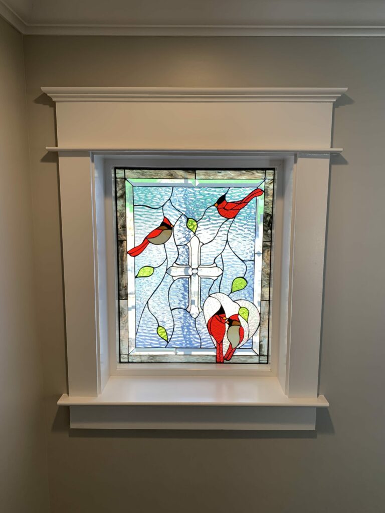 A rectangular leaded stained glass window featuring a prominent white cross. Three vibrant red cardinal birds are depicted: two are flying near the top, and one is nesting or perching at the bottom right. Green leaves are scattered throughout the design, set against a textured light blue background resembling water or sky. The panel is bordered by clear beveled and mottled grey glass pieces.