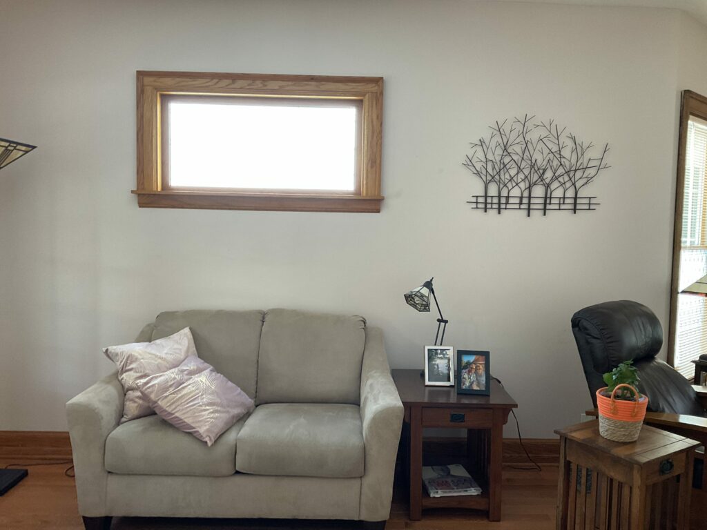A horizontal rectangular window with a light wooden frame, positioned above a beige two-seater sofa and an end table in a modern living room. The window is bright with natural light.