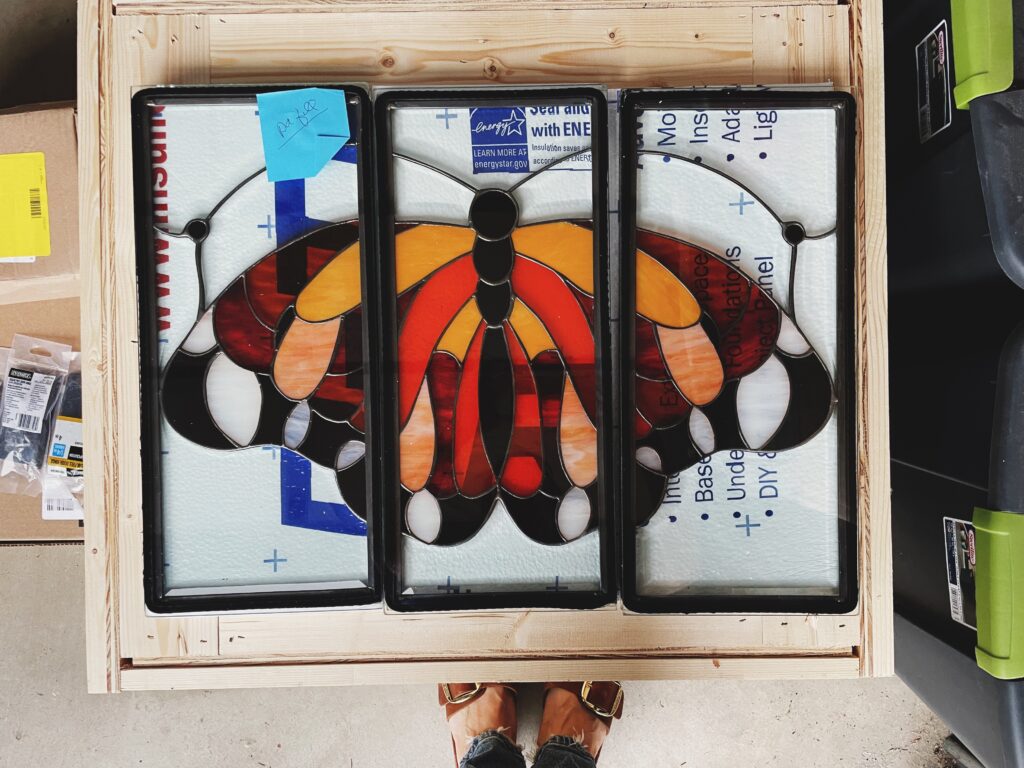 A three-panel stained glass artwork depicting a monarch butterfly with wings spread, viewed from above. The butterfly's body is black, and its wings feature vibrant shades of orange, red, and yellow swirled glass, accented with clear textured and opaque white sections, all outlined with dark lead lines. Each rectangular panel is framed in dark metal and rests within a light wooden shipping crate, with white insulating material visible behind the glass.