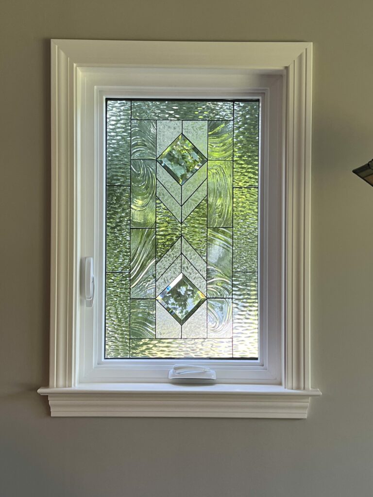 A rectangular clear leaded stained glass window panel featuring a geometric design with central beveled diamonds and surrounding clear textured glass, set within a white window frame with decorative trim. Green foliage is visible outside through the glass.