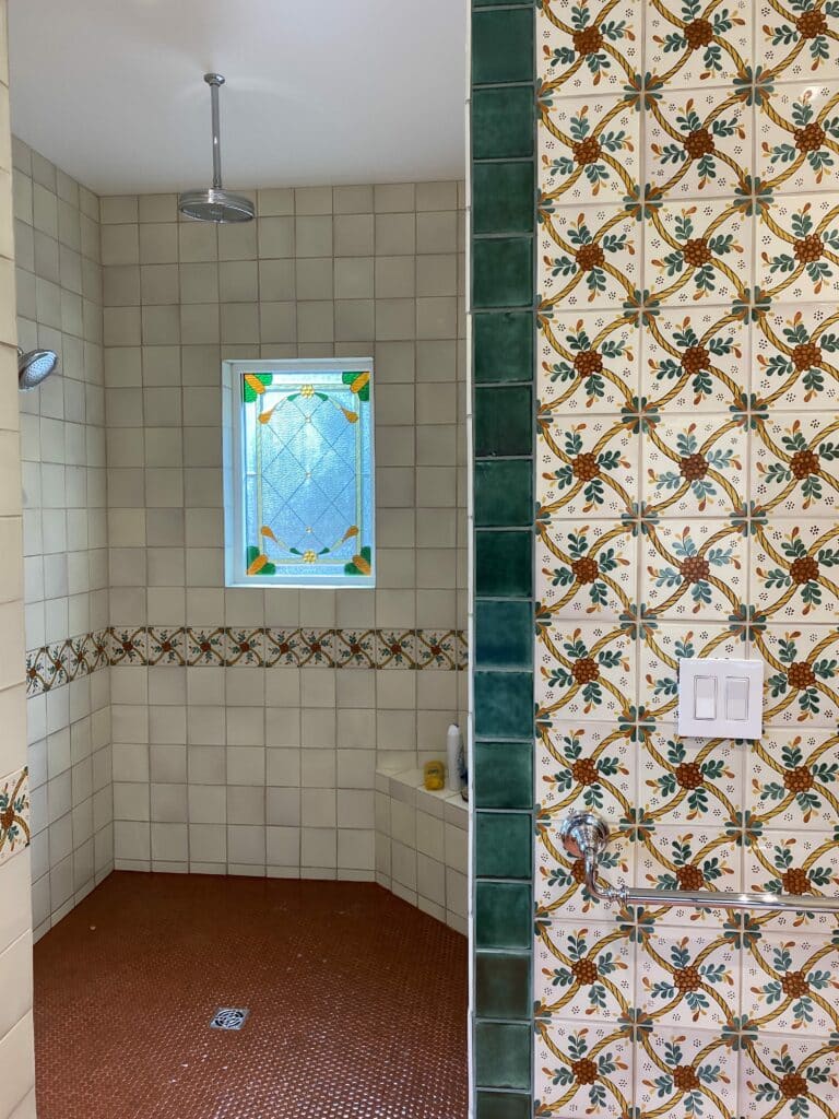 A small rectangular leaded stained glass window featuring clear textured glass and an Art Nouveau-inspired border with yellow, green, and orange floral elements, set into a custom-tiled shower wall.
