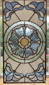 A rectangular stained glass panel featuring a central white and yellow stylized flower medallion, surrounded by a textured green ring, with blue floral motifs in the corners.