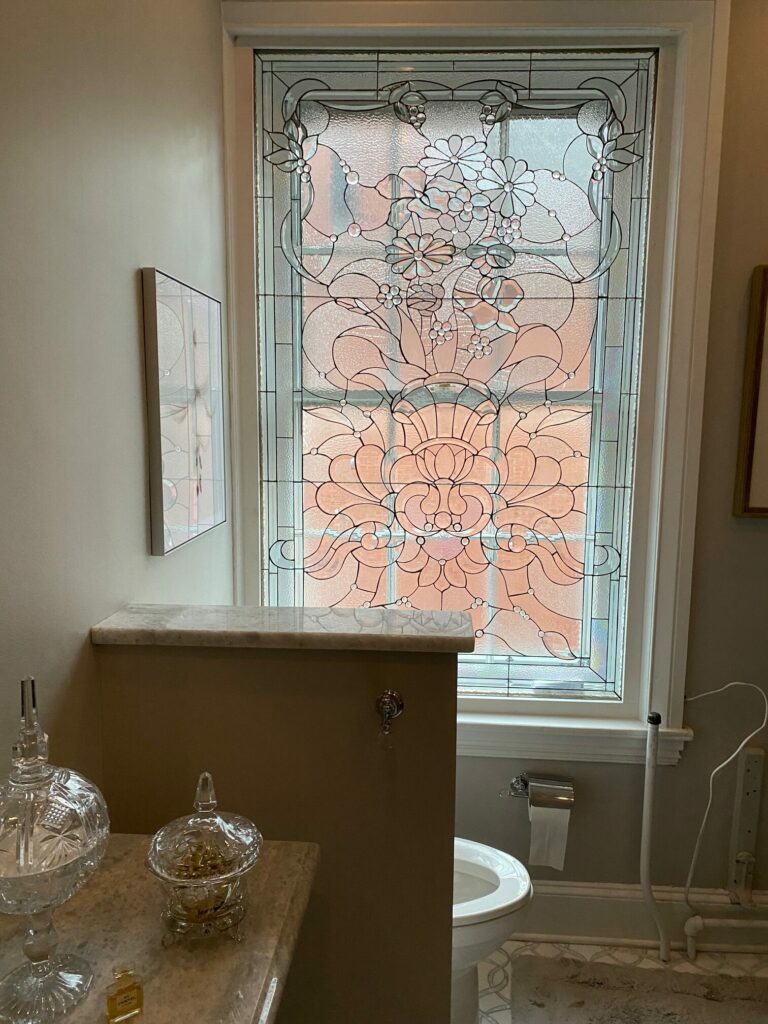 A large, rectangular clear leaded stained glass window panel installed in a bathroom, featuring an intricate Victorian floral design with scrolling vines, multiple clear textured glass types, and small round beveled accents, providing privacy.