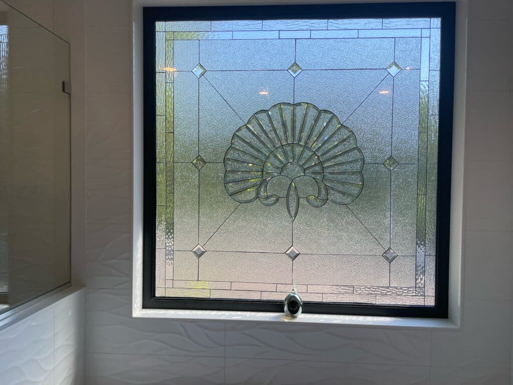 A rectangular leaded stained glass window with a black frame, featuring a central fan or shell-shaped medallion made of clear, heavily textured, and subtly beveled glass. The design includes a geometric grid with clear diamond bevels at the intersections, and additional clear textured glass patterns in the borders, providing privacy while allowing light. The window is installed in a modern bathroom with white wavy wall tiles.