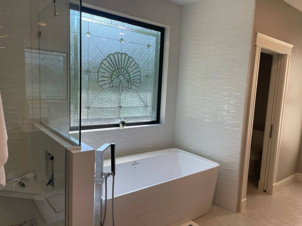 A rectangular leaded glass window featuring a prominent central fan-shaped medallion crafted from clear beveled and textured glass. The surrounding glass panels are clear textured with small diamond bevel accents, all set within a black window frame above a white freestanding bathtub in a modern bathroom with white wavy tiled walls.