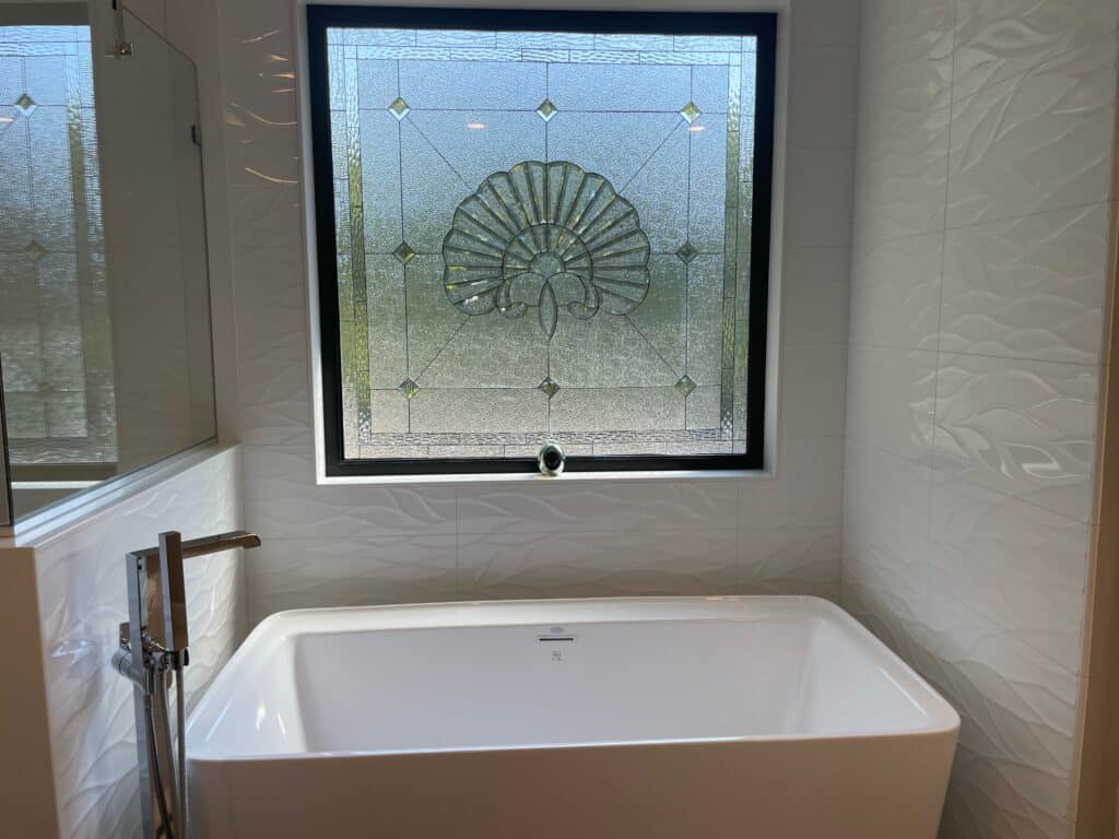 A rectangular, black-framed leaded stained glass window featuring a prominent central shell or fan-like motif crafted from clear, textured, and beveled glass. The motif is surrounded by clear textured glass panels and small beveled diamonds. The window is positioned above a white freestanding bathtub in a modern bathroom with white textured wave-patterned wall tiles.